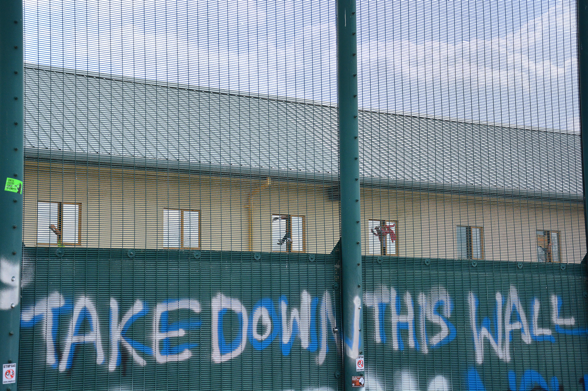 Photo of the fence outside Yarl's Wood with graffiti saying "Take Down This Wall" and people detained inside putting their arms out the window in the background.